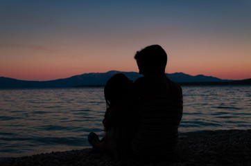 Mother holding her baby girl in her lap, sitting on a beach after sunset.