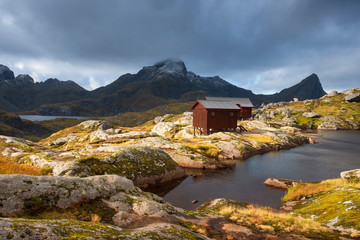 Lofoten Islands Norway. Mountain autumn landscape. Hike to Mount Munkan, wooden houses, a shelter and a lake in the sunlight against a stormy sky - obrazy, fototapety, plakaty