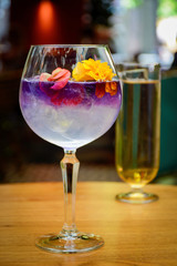 Cocktail glasses. Ice cold modern craft cocktail garnished by some flowers on a table with blurry restaurant background.