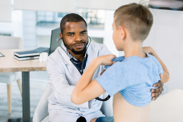 African male doctor and child boy patient. Smiling pediatrician man sees boy in medical office. African American male pediatrician with stethoscope listening to lung and heart sound of little boy