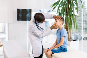 Obraz premium Young African American male doctor examines the eyes of his patient little boy, sitting on the couch in medical office