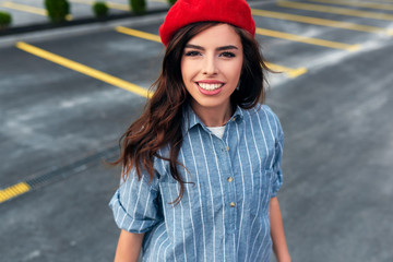 Portrait of beautiful brunette young woman smiling broadly with toothy smile with red lips, wearing blue shirt and red beret hat, looking to the camera, posing on the city.