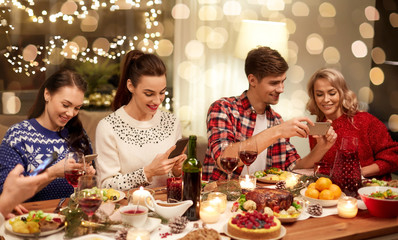 holidays and celebration concept - happy friends with smartphones having christmas dinner at home