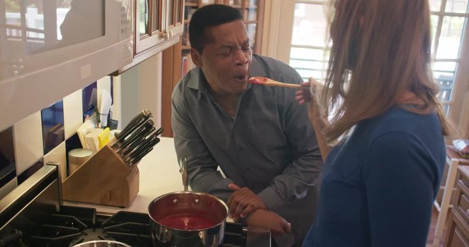 Mature Caucasian Woman Feeding Home Made Marinara Sauce To Husband In Kitchen. African American Man Tasting His Wifes Cooking And Smiling. Slow Motion 4k Handheld