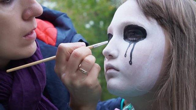 Mom painting daughter face for Halloween party in the courtyard of his house. Children's Zombie Makeup. Halloween and day of the dead concept.