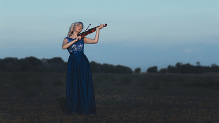 beautiful young woman in long dress playing violin in field, girl figure on horizon on nature...
