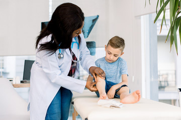 African woman doctor in a white coat carefully provides first aid. She examines the boy leg. The kid patiently observes the process, sitting on the couch.