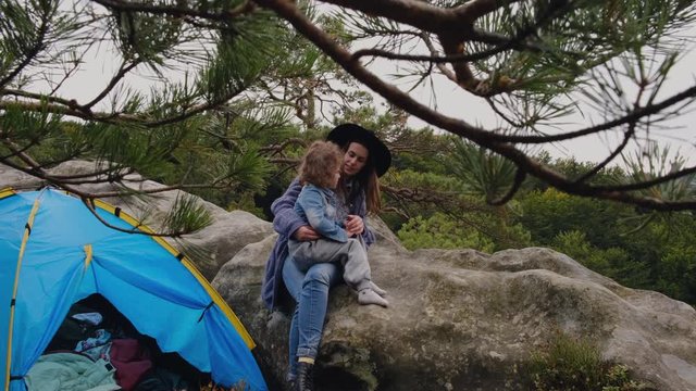 A young mother with a small daughter looks afar on the top of mountains. Mother with daughter sitting near to blue tent in wild national park forest. People and nature concept