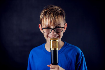 A portrait of a boy with eyeglasses holds microphone in front of dark background. Children and entertainment concept