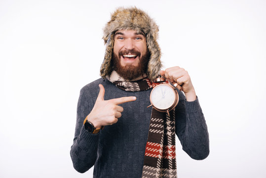 Look at the clock Bearded man pointing at an alarm clock, wearning warm clothing, smiling at the camera.