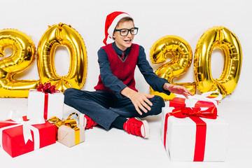 Happy little boy in Santa hat, stylish baby with Christmas gifts, sits on white background with...