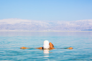 mainstream travel photography from Israeli health care resort girl in a white hat back to camera floating on calm smooth dead sea salty water surface, copy space  