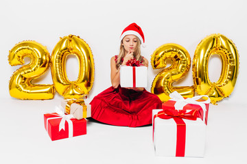Pensive little girl in Santa hat, stylish baby sitting pensive with Christmas gifts sitting on...