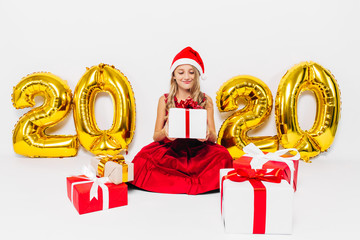 Happy little girl in Santa hat, stylish baby sitting with Christmas gifts sitting on white...