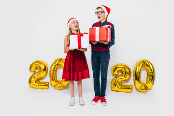Happy little girl and boy wearing Santa hat, stylish brother and sister are rejoicing holding...