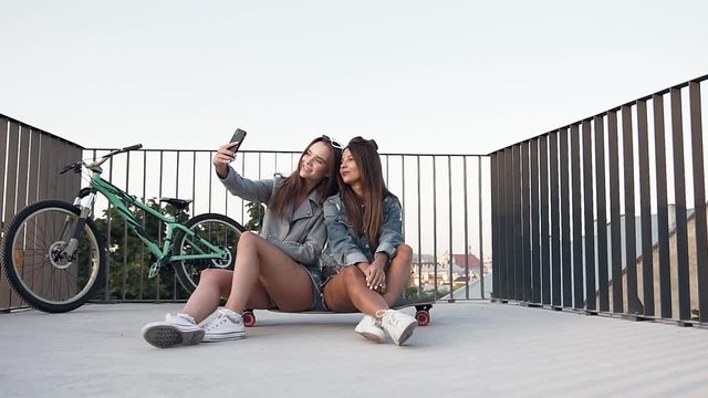 Pretty jovial sexy girls sitting on the skateboard and making selfie with funny faces on the roof terrace