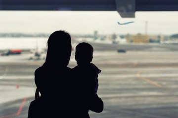 Silhouette of mother holding on hands little toddler boy with window of airport on background....