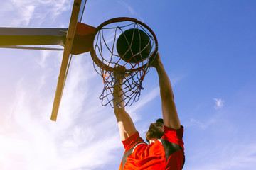 Bottom view of an athlete performing a dunk in a basketball hoop in a jump on a background of blue sky and bright sun. Sports lifestyle, desire for victory and success. Young guy basketball player.
