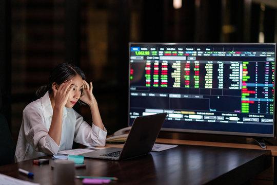 Asian businesswoman holding hands on head and sitting at the desk work with computer and stock exchange monitor in night worklate office. Stress female headache,tired and burnout with hard work.