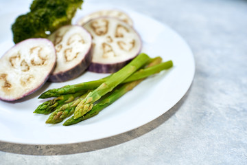 Healthy eating concept. Steamed vegetables in a white plate on a blue table. Eggplant, broccoli, asparagus.