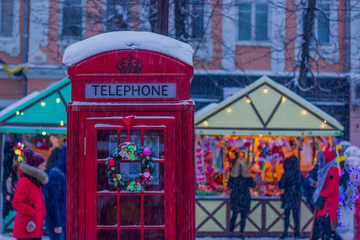 winter holidays and Christmas fair season celebration concept picture of red phone box foreground and shopping houses with unfocused people background in snowing weather time 