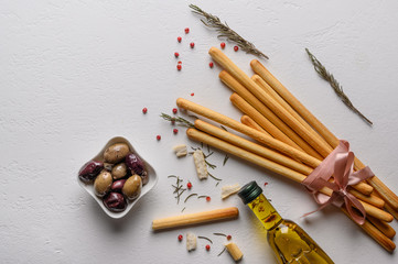 Traditional Italian breadsticks grissini lie on a light textured table tied with tape. Next to a vase of olives and a bottle of olive oil