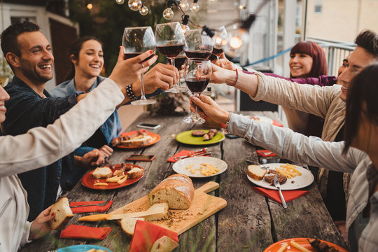 Group of young people having lunch on a terrace of an apartment at sunset - Millennials having fun together on a day of celebration - Toast with glasses of red wine