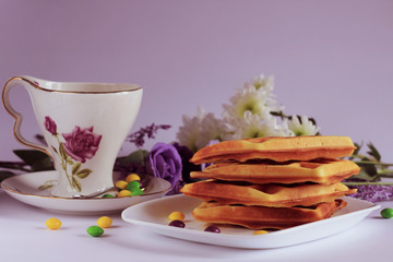 Natural crispy waffles with which lie on a white plate. Morning breakfast with a cup of tea and colored sweets on a light background in lilac colors. Bluer effect