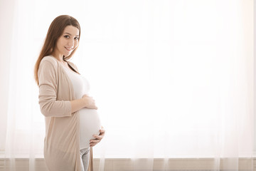 Young pregnant woman embracing her belly, standing near window - obrazy, fototapety, plakaty