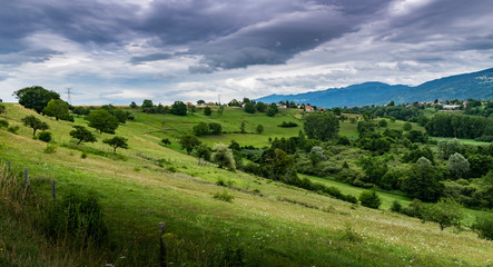 Fototapeta premium View of the mountain landscape,green pastures, trees, forests on the slopes of hills and farmhouses with a cloudy sky in the background.Brens,Haute-Savoie in France.