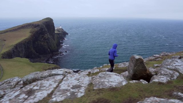 Tracking follow shot of a girl walking on the edge with cliffs in foreground and Neist Point Lighthouse and Atlantic Ocean in the background trying to take a photograph in Scotland, Isle of Skye