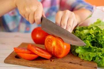 Slice Tomatoes in Half. Red tomato on the chopping Board, in the kitchen. Female Chef cuts one red tomato with a knife on wooden cutting board. The process of cooking, close-up.