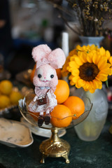 Obraz premium Cute bunny doll. Kawaii bunny felt doll sitting on top of piles of oranges and sun flowers on the background. Cafe hopping and toy photography concept.