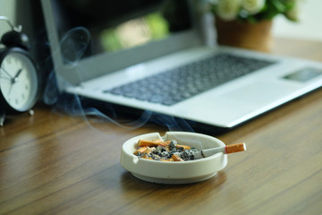 Many cigarette in white ashtray on the desk with laptop computer. Smoking to relieve stress at work but unhealthy