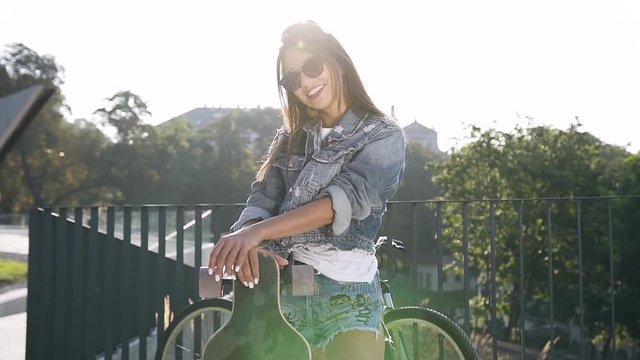 Beautiful flirtatious young woman with sunglasses and tempting jeans clothes standing with skateboard and posing on camera on the background of city landscape
