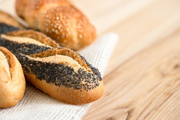 Poppy seed baguette and bread rolls lying on white burlap on light wood. Space for text.