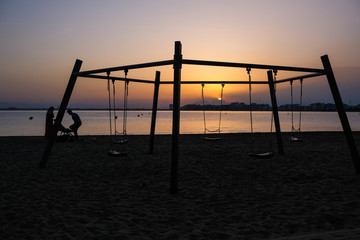 Children's swing on the beach on a background of sunset and sea. Silhouettes of a family of two parents and a child. Concept of family vacation. Summer sunset in Spain, Catalunya. Playground on beach.