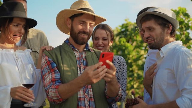 Funny farmer showing his friends disgusting pictures on smartphone. Group of multi-ethnic people with flinched faces staying in vineyard.