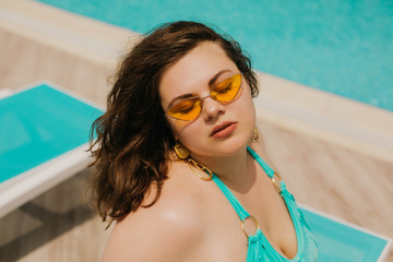Attractive busty curvy woman in yellow sunglasses and a blue swimsuit resting by the pool. Stylish accessories, fringe, fashion for plus size. Bodypositive, natural real beauty, resort, summer