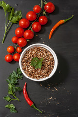 Cooked buckwheat porridge in a deep plate on a black wooden background with vegetables. Copy space. Top view