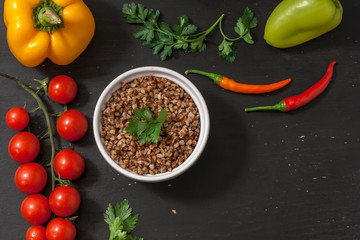 Cooked buckwheat porridge in a deep plate on a black wooden background with vegetables. Copy space. Top view