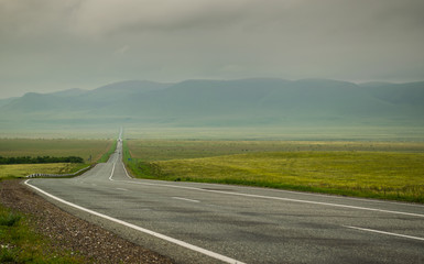 Highway road in Khakassia with green grass steppe and sayan mountains in fog and gray clouds in summer day, Siberia, Russia