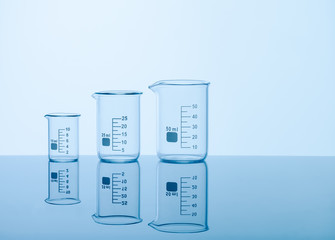 Three empty measuring beakers sitting on a mirror blue surface, glass lab containers standing in gradual order on a table, transparent light through laboratory flasks, precise comparing measurements 