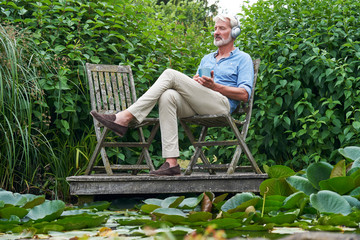 Mature Man Relaxing In Garden Listening To Music On Wireless Headphones On Jetty By Lake