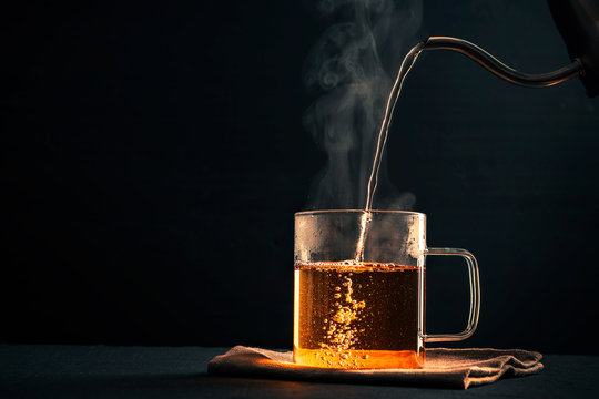The process of brewing tea, pouring hot water from the kettle into the Cup,  steam coming out of the mug, water droplets on the glass, black background  Stock Photo