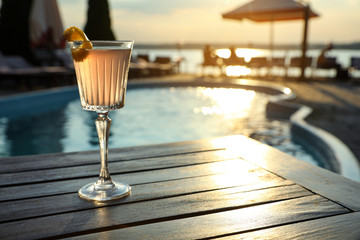 Glass of fresh summer cocktail on wooden table near swimming pool outdoors at sunset. Space for text