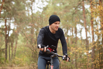 Fototapeta na wymiar Horizontal shot of young handsome guy riding bike in forest, man wearing black sportwearand cap, male looks concentrated, looking down, enjoys spending time in open air. Healthy lifestyle concept.