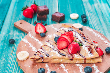 Crepe with Strawberries , Berries and Bananas and Brownies - Strawberry Crepe with Fruits with Chocolate Sauce on Top and Sugar Powder on Blue Wood Background