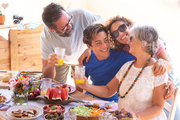 Cheerful family, three generations, hugging each other eating vegan food together on the wooden table. Senior woman with son, daughter-in-law and nephew. Bright sunlight.
