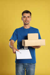 Happy young courier with parcels and clipboard on yellow background